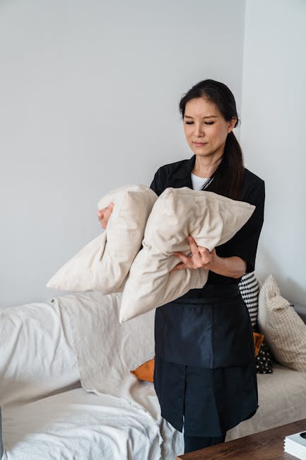 A person wearing a dark jacket is using a handheld vacuum cleaner to thoroughly clean a yellow and white patterned cushion placed on a dark, fabric-upholstered sofa in a well-lit room. The surface of 